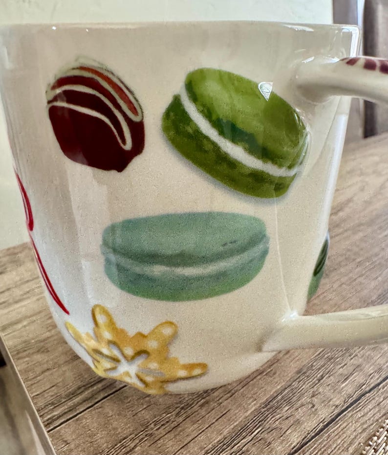 May include: Cream ceramic mug decorated with illustrations of a chocolate truffle, green and blue macarons, and a gold snowflake. The mug has a curved handle and is set on a wooden surface.
