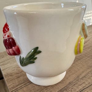 K&ouml;nnte beinhalten: Ein wei&szlig;er Keramik-Blumentopf mit rundem K&ouml;rper und kleinem Sockel. Der Topf ist mit farbenfrohen, erhabenen Reliefdesigns verziert, darunter ein rotes und rosa Blumenelement und gr&uuml;ne Bl&auml;tter.