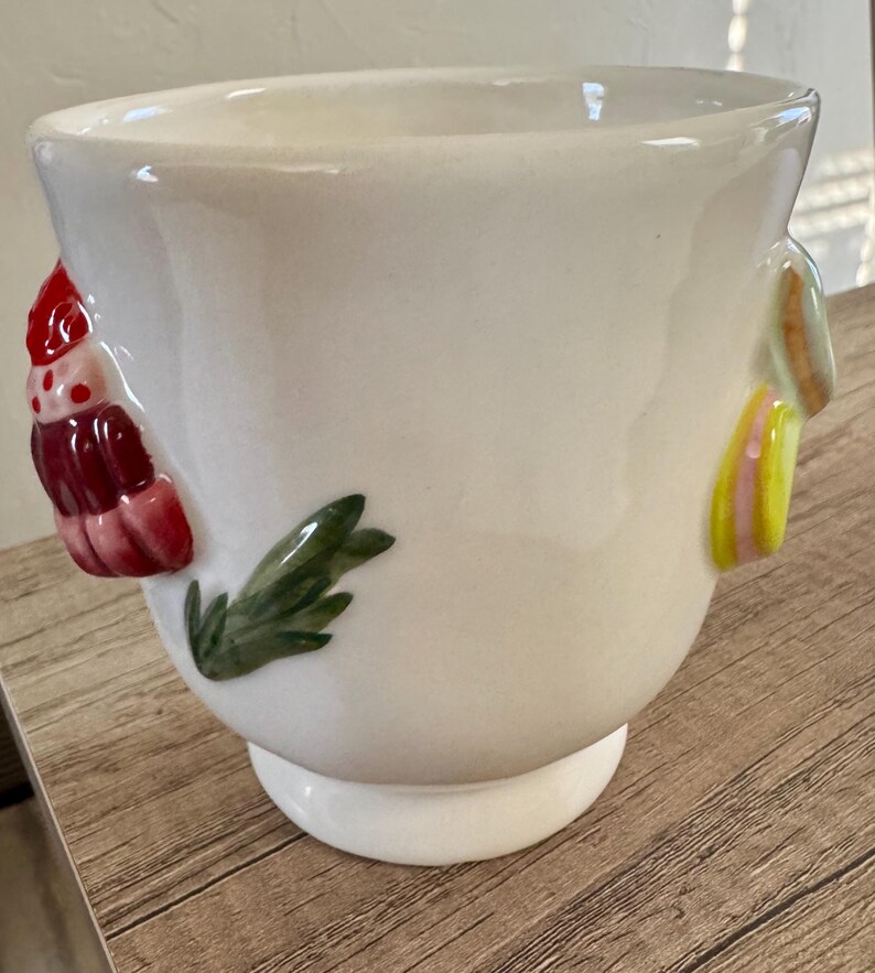 K&ouml;nnte beinhalten: Ein wei&szlig;er Keramik-Blumentopf mit rundem K&ouml;rper und kleinem Sockel. Der Topf ist mit farbenfrohen, erhabenen Reliefdesigns verziert, darunter ein rotes und rosa Blumenelement und gr&uuml;ne Bl&auml;tter.