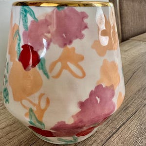May include: A white ceramic vase with a gold rim, decorated with watercolour floral patterns in pink, orange, and red. The vase has a gently curved shape and is suitable for displaying flowers or as a decorative piece.