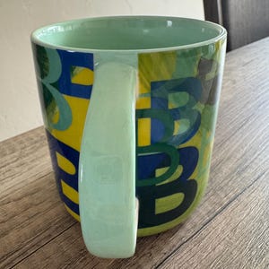 May include: A green ceramic mug with a yellow, blue, and green abstract design. The mug has a large handle.