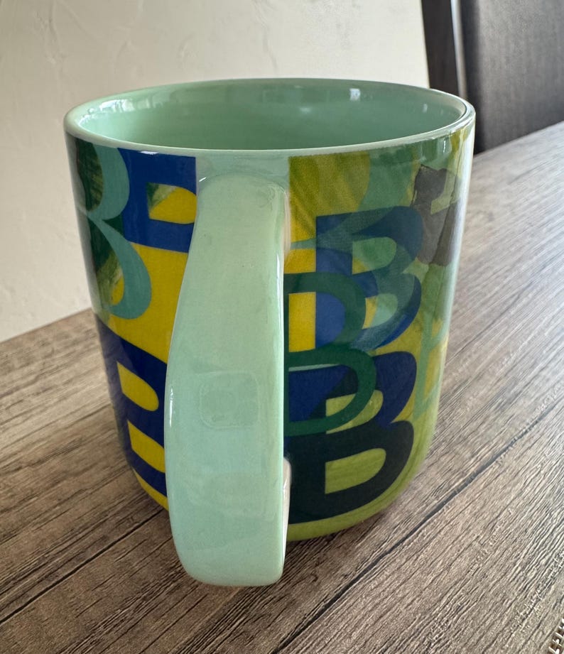 May include: A green ceramic mug with a yellow, blue, and green abstract design. The mug has a large handle.
