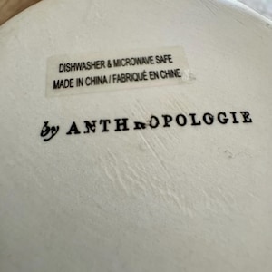 May include: Close-up of a white ceramic surface with the text "DISHWASHER & MICROWAVE SAFE" and "MADE IN CHINA / FABRIQU&Eacute; EN CHINE". The words "by ANTHROPOLOGIE" are printed below.