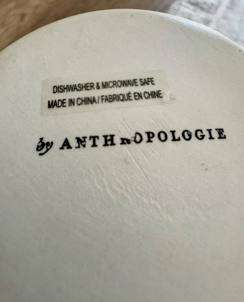 May include: Close-up of a white ceramic surface with the text "DISHWASHER & MICROWAVE SAFE" and "MADE IN CHINA / FABRIQU&Eacute; EN CHINE". The words "by ANTHROPOLOGIE" are printed below.