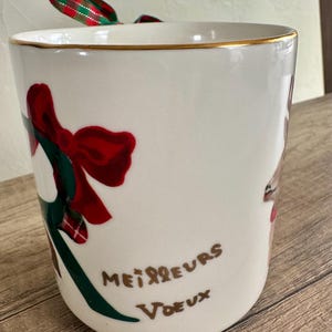 Puede incluir: Una taza de cer&aacute;mica blanca con borde dorado, con un lazo rojo y detalles verdes. La taza tiene las palabras "Meilleurs Voeux" en escritura marr&oacute;n. Una cinta roja, verde y blanca est&aacute; atada a la parte superior.