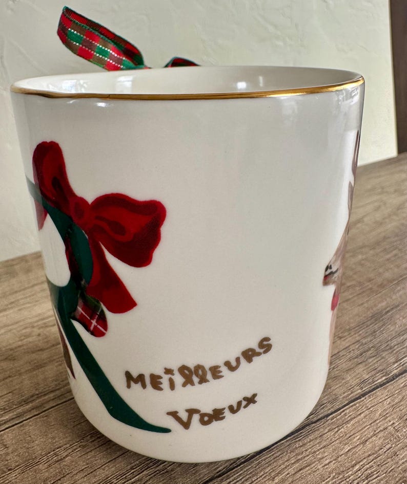 Puede incluir: Una taza de cer&aacute;mica blanca con borde dorado, con un lazo rojo y detalles verdes. La taza tiene las palabras "Meilleurs Voeux" en escritura marr&oacute;n. Una cinta roja, verde y blanca est&aacute; atada a la parte superior.