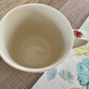 May include: Cream ceramic mug with a red and white peppermint candy design on the handle. The mug is empty and rests on a patterned placemat and floral cloth.