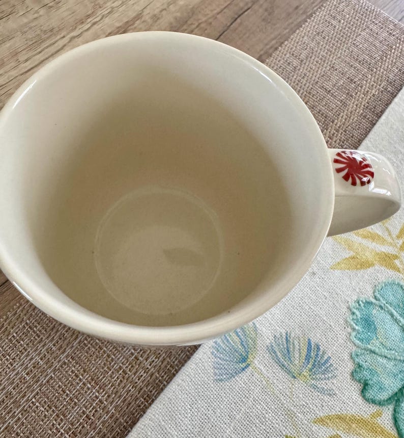 May include: Cream ceramic mug with a red and white peppermint candy design on the handle. The mug is empty and rests on a patterned placemat and floral cloth.