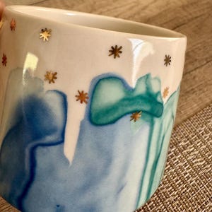 May include: A ceramic mug featuring a white upper section decorated with small gold star accents. The lower portion displays a fluid, watercolor-style pattern in shades of blue and teal, creating a smooth transition of colors across the surface.