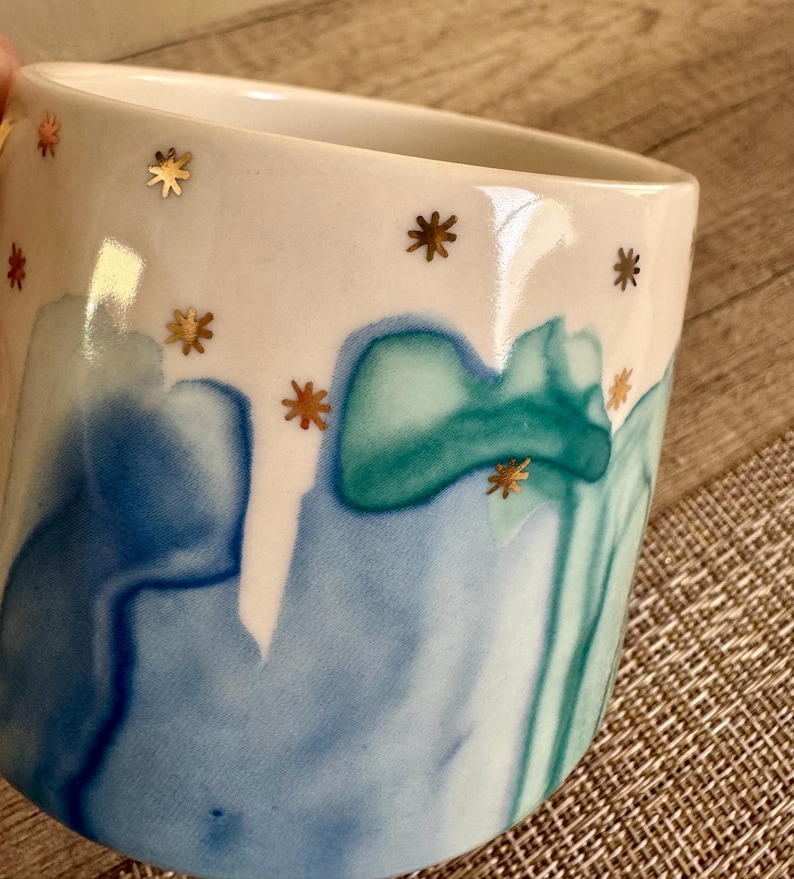 May include: A ceramic mug featuring a white upper section decorated with small gold star accents. The lower portion displays a fluid, watercolor-style pattern in shades of blue and teal, creating a smooth transition of colors across the surface.