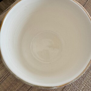 Pode incluir: Uma caneca de cer&acirc;mica creme com uma abertura larga e redonda e uma al&ccedil;a. A borda da caneca tem uma fina faixa dourada. A caneca est&aacute; sobre uma superf&iacute;cie de madeira.