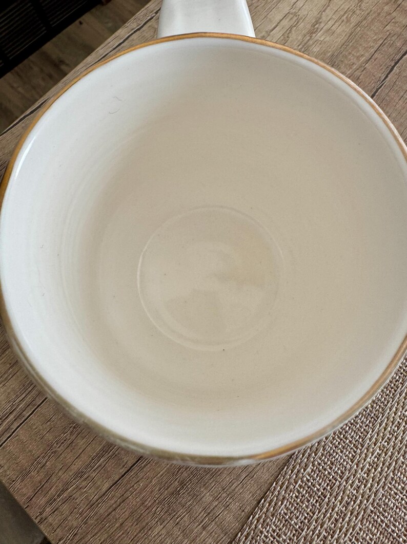 Pode incluir: Uma caneca de cer&acirc;mica creme com uma abertura larga e redonda e uma al&ccedil;a. A borda da caneca tem uma fina faixa dourada. A caneca est&aacute; sobre uma superf&iacute;cie de madeira.