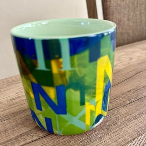 May include: A ceramic mug with a light green interior and a colorful exterior. The exterior features a design of overlapping blue, yellow, and green letters. The mug sits on a wooden surface.