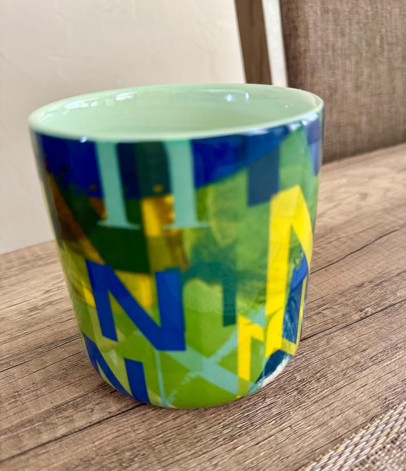 May include: A ceramic mug with a light green interior and a colorful exterior. The exterior features a design of overlapping blue, yellow, and green letters. The mug sits on a wooden surface.