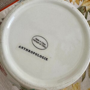 May include: A white ceramic vase with the word "ANTHROPOLOGIE" printed in black. The base of the vase has a sticker that says "Made in China Fabriqu&eacute; en Chine". The vase is on a floral patterned surface.