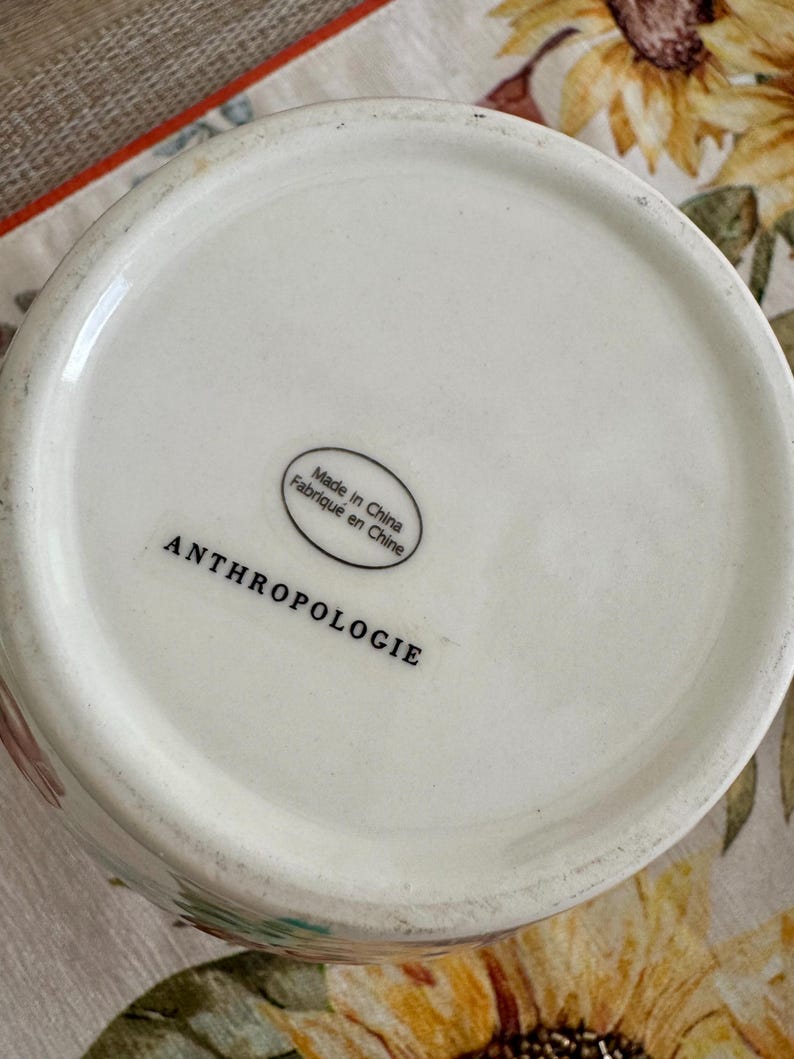 May include: A white ceramic vase with the word "ANTHROPOLOGIE" printed in black. The base of the vase has a sticker that says "Made in China Fabriqu&eacute; en Chine". The vase is on a floral patterned surface.