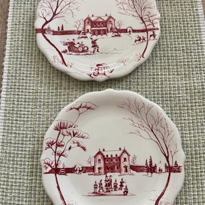 May include: Two cream-colored ceramic plates with scalloped edges, each featuring a detailed maroon-colored winter scene. The top plate depicts a horse-drawn sleigh, while the bottom plate shows a group of figures standing before a large house.