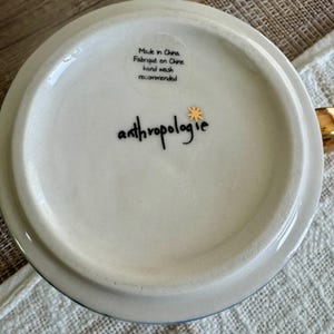 May include: The white underside of a ceramic mug featuring the black text anthropologie with a small gold star icon. A clear sticker displays the text Made in China, Fabriqu&eacute; en Chine, and hand wash recommended in black lettering.