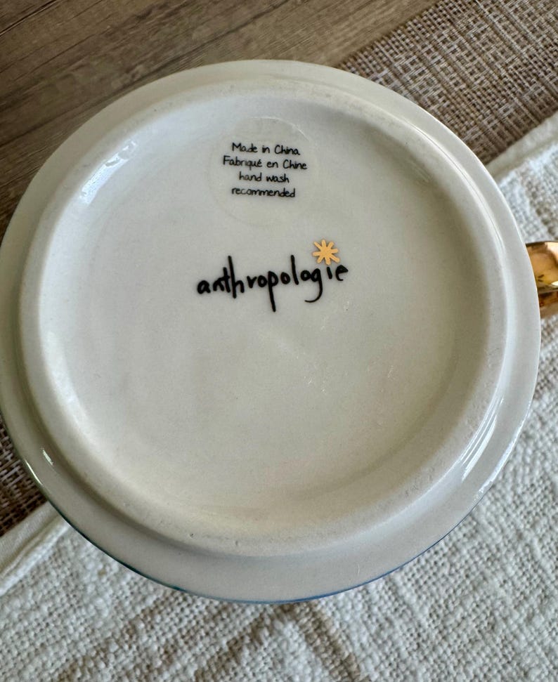 May include: The white underside of a ceramic mug featuring the black text anthropologie with a small gold star icon. A clear sticker displays the text Made in China, Fabriqu&eacute; en Chine, and hand wash recommended in black lettering.