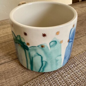 May include: A ceramic mug with a white interior and exterior featuring a painted design of abstract teal and blue watercolor-style vertical streaks. The upper portion of the mug is decorated with small, metallic gold star-shaped accents.