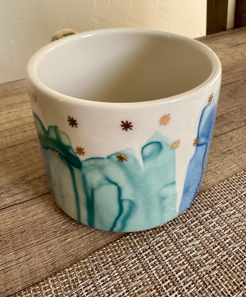 May include: A ceramic mug with a white interior and exterior featuring a painted design of abstract teal and blue watercolor-style vertical streaks. The upper portion of the mug is decorated with small, metallic gold star-shaped accents.
