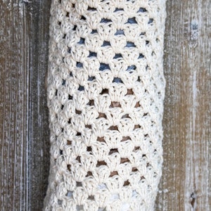 May include: A white crocheted bag for storing plastic grocery bags. The bag is hanging on a wooden wall. The bag is made with a simple crochet stitch and has a large opening at the top.