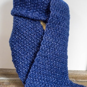 May include: A blue crocheted scarf with a textured pattern. The scarf is folded and draped over a wooden surface.