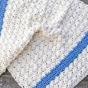 May include: A crocheted blanket with a blue stripe down the center. The blanket is made with a textured stitch and is a light cream color.