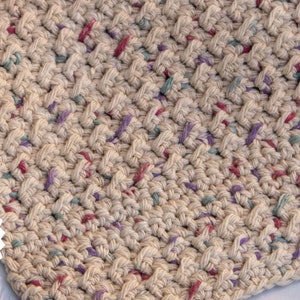 May include: A close-up of a beige crochet dishcloth with a textured pattern. The dishcloth has a speckled appearance with small flecks of pink, purple, and blue yarn.