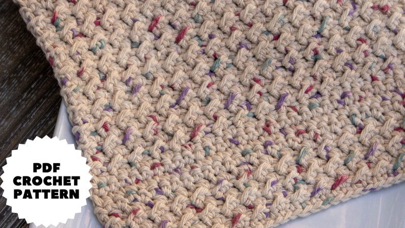 May include: A close-up of a beige crochet dishcloth with a textured pattern. The dishcloth has a speckled appearance with small flecks of pink, purple, and blue yarn.