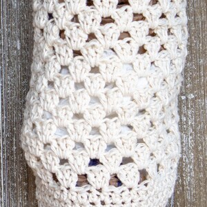 May include: A crocheted bag made with white yarn, designed to hold plastic grocery bags. The bag is hanging on a wooden surface.