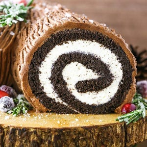 Peut inclure: Bûche de Noël au chocolat avec une garniture de crème blanche, présentée sur une planche de service en bois. Le gâteau est décoré de glaçage au chocolat, de canneberges sucrées et de brins de verdure, parfait pour les fêtes.