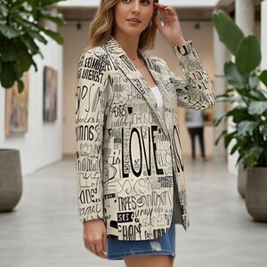 Puede incluir: Blazer color crema con texto negro y la palabra "LOVE" en un lugar destacado. El blazer tiene solapas con muescas y mangas largas. La modelo lleva un top blanco y pantalones cortos vaqueros.