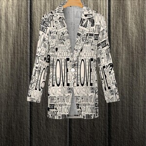 Puede incluir: Una blazer blanca con un dise&ntilde;o de palabras en negro, incluyendo la palabra "LOVE" en un lugar destacado. La blazer est&aacute; colgada en una percha de madera sobre un fondo de madera texturizada. El dise&ntilde;o incluye palabras como "SUNRISE" y "CHOCOLATE".
