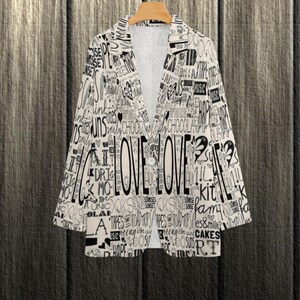 Puede incluir: Blazer color crema con un dise&ntilde;o de texto negro, incluyendo la palabra "LOVE" prominentemente mostrada. El blazer est&aacute; abotonado y colgado en una percha de madera sobre un fondo de vetas oscuras. El dise&ntilde;o de texto incluye palabras como "cakes" y "music".