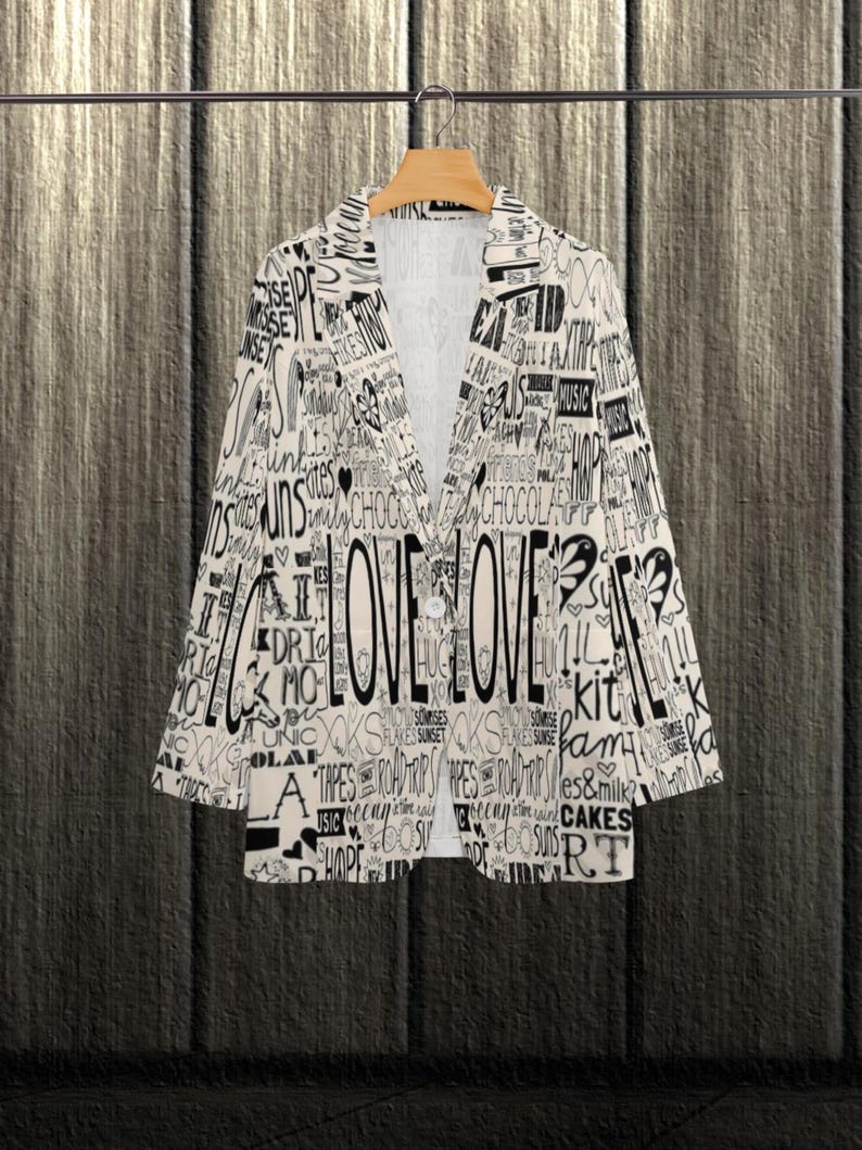 Puede incluir: Blazer color crema con un dise&ntilde;o de texto negro, incluyendo la palabra "LOVE" prominentemente mostrada. El blazer est&aacute; abotonado y colgado en una percha de madera sobre un fondo de vetas oscuras. El dise&ntilde;o de texto incluye palabras como "cakes" y "music".