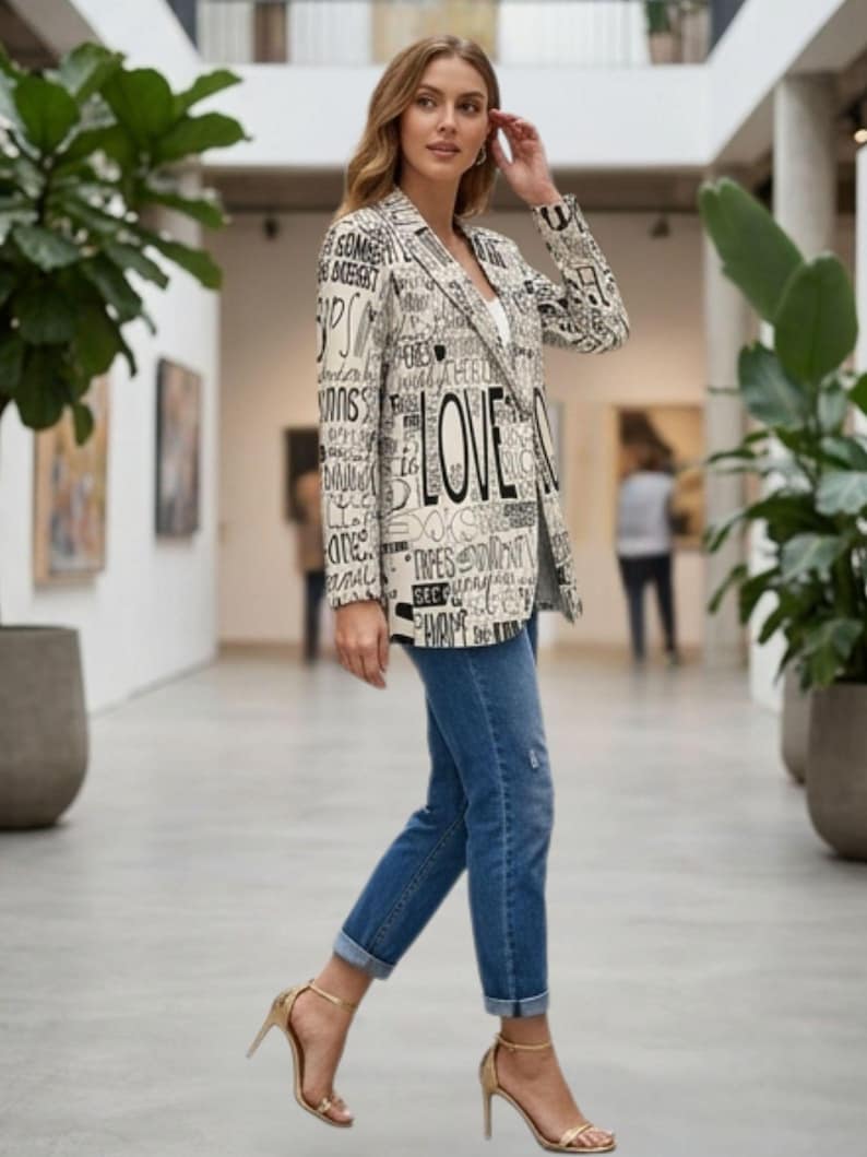 Puede incluir: Una blazer crema con texto negro, incluyendo la palabra "LOVE", combinada con vaqueros azules y tacones dorados. El conjunto se luce en un pasillo luminoso con plantas.
