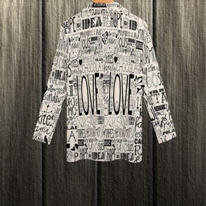 Puede incluir: Una blazer blanca con un dise&ntilde;o de texto negro, que incluye la palabra "LOVE" de forma destacada. La chaqueta de manga larga est&aacute; colgada en una percha de madera sobre un fondo texturizado. El dise&ntilde;o incluye palabras como "idea", "esperanza" y "cometas".