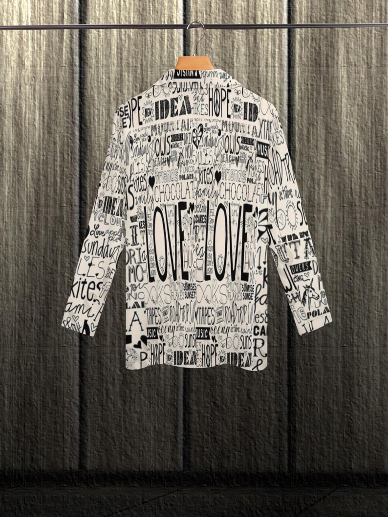 Puede incluir: Una blazer blanca con un dise&ntilde;o de texto negro, que incluye la palabra "LOVE" de forma destacada. La chaqueta de manga larga est&aacute; colgada en una percha de madera sobre un fondo texturizado. El dise&ntilde;o incluye palabras como "idea", "esperanza" y "cometas".