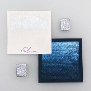 Könnte beinhalten: Zwei kleine silberne Metallic-Farbquadrate und ein größeres Quadrat aus dunkelblauem Metallic-Farb. Das größere Quadrat ist schwarz gerahmt und trägt den Schriftzug "Glam" in Lila.