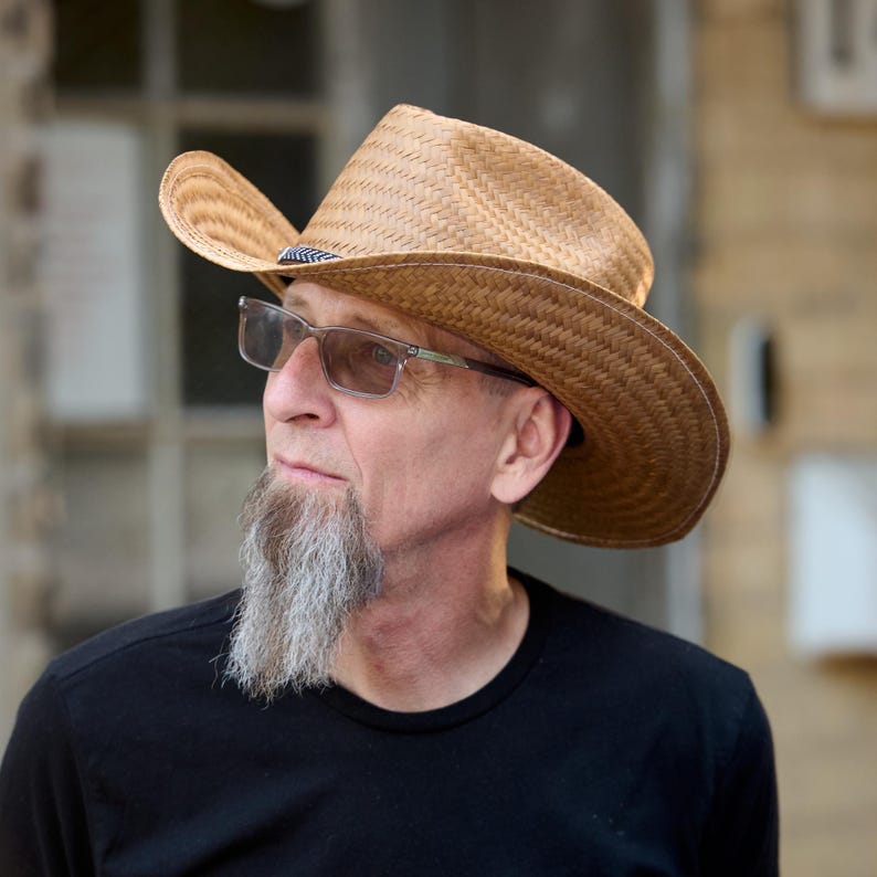 May include: A man with a gray beard and glasses is wearing a brown straw cowboy hat. He is wearing a black t-shirt.