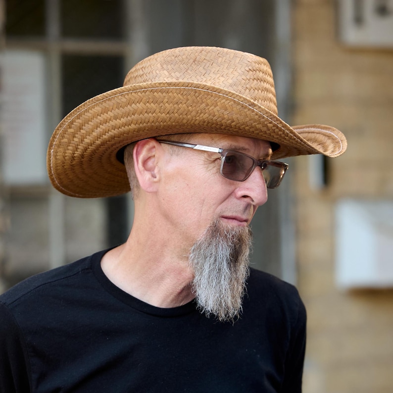 May include: A man with a gray goatee wearing a brown straw cowboy hat and brown-framed glasses.