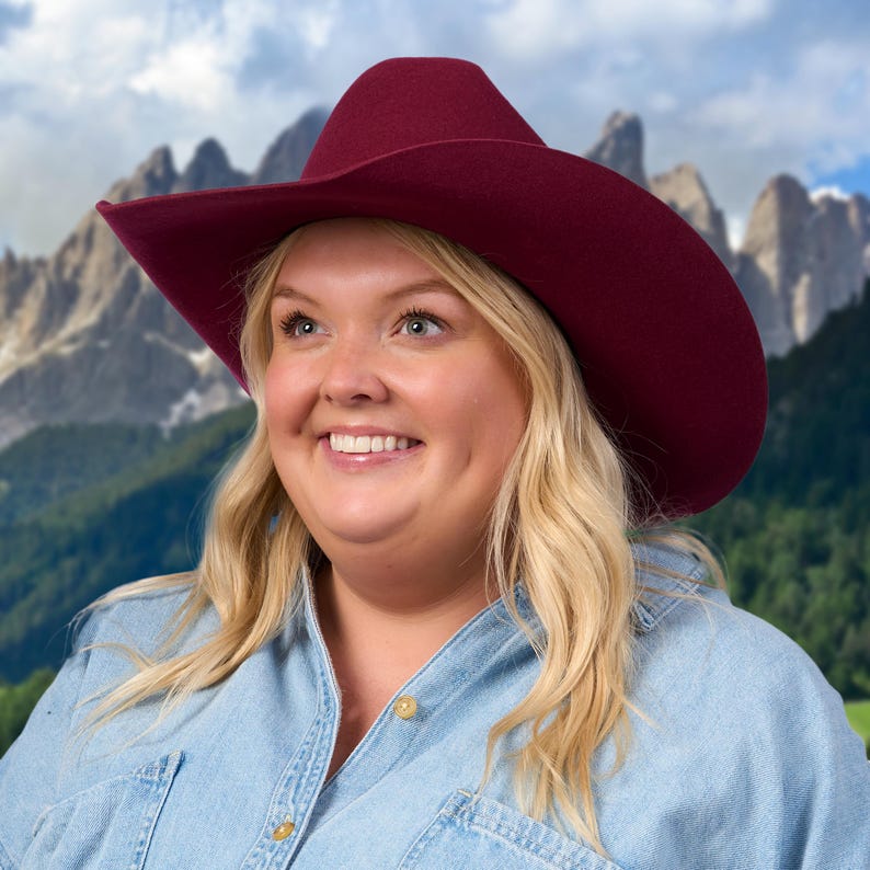 K&ouml;nnte beinhalten: Eine Frau mit blonden Haaren tr&auml;gt einen burgunderfarbenen Cowboyhut und ein hellblaues Jeanshemd. Sie l&auml;chelt und schaut nach oben. Der Hintergrund ist ein verschwommenes Bild von Bergen.