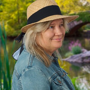 May include: A woman wearing a straw hat with a black ribbon and a denim jacket. The hat has a black ribbon tied around it and hangs down the side of her neck. She is looking to the right of the frame.