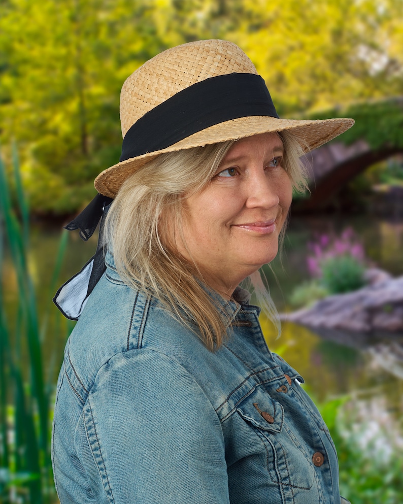 May include: A woman wearing a straw hat with a black ribbon and a denim jacket. The hat has a black ribbon tied around it and hangs down the side of her neck. She is looking to the right of the frame.