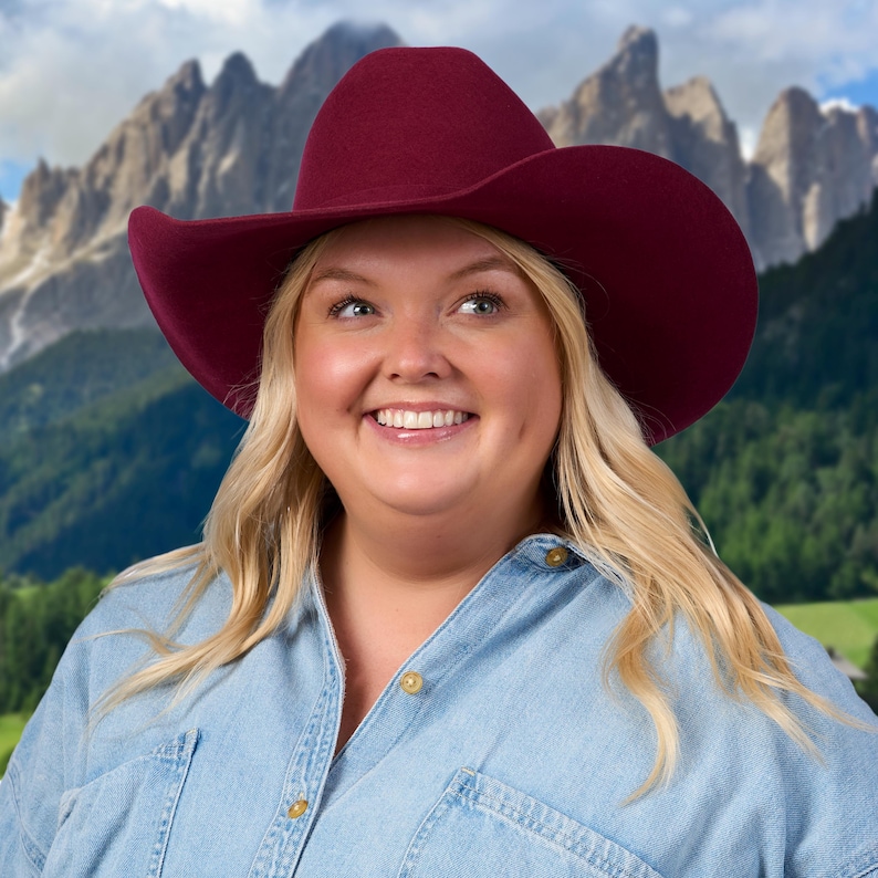 K&ouml;nnte beinhalten: Eine Frau mit blonden Haaren tr&auml;gt ein Jeanshemd und einen burgunderfarbenen Cowboyhut. Sie l&auml;chelt und schaut nach oben. Der Hintergrund ist ein verschwommenes Bild von Bergen und B&auml;umen.