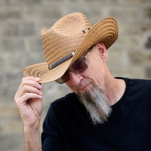 May include: A man wearing a brown straw cowboy hat with a black band and a black t-shirt. He has a gray beard and is looking down.