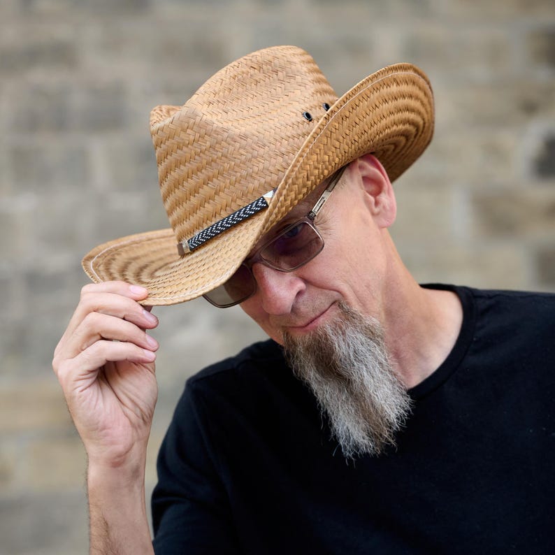 May include: A man wearing a brown straw cowboy hat with a black band and a black t-shirt. He has a gray beard and is looking down.