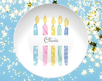 May include: White ceramic plate with a watercolor design of birthday candles in blue, pink, yellow, and green. The name "Olivia" is written in cursive. The plate is decorated with gold star confetti on a light blue background.