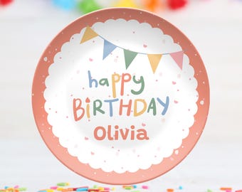 May include: A round, peach-colored plate with a white center featuring the words "happy BIRTHDAY Olivia" in colorful letters. The plate has a scalloped edge and a banner design with colorful flags. Sprinkles are scattered around the plate.